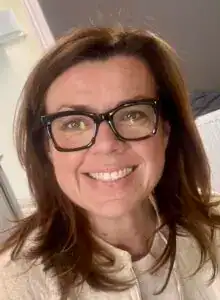 A woman with brown hair and glasses smiling indoors at Shine At The Hollies.