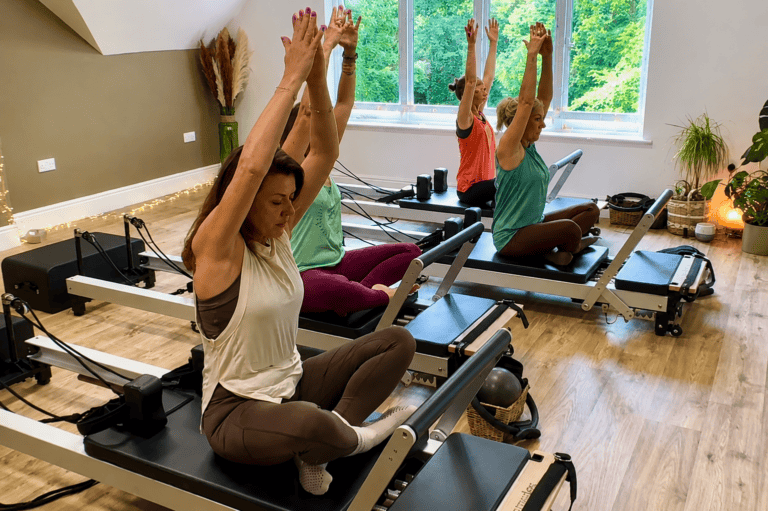 Group of women sitting on reformer Pilates machines doing an over head stretch during a Reformer Foundations class at Shine At The Hollies