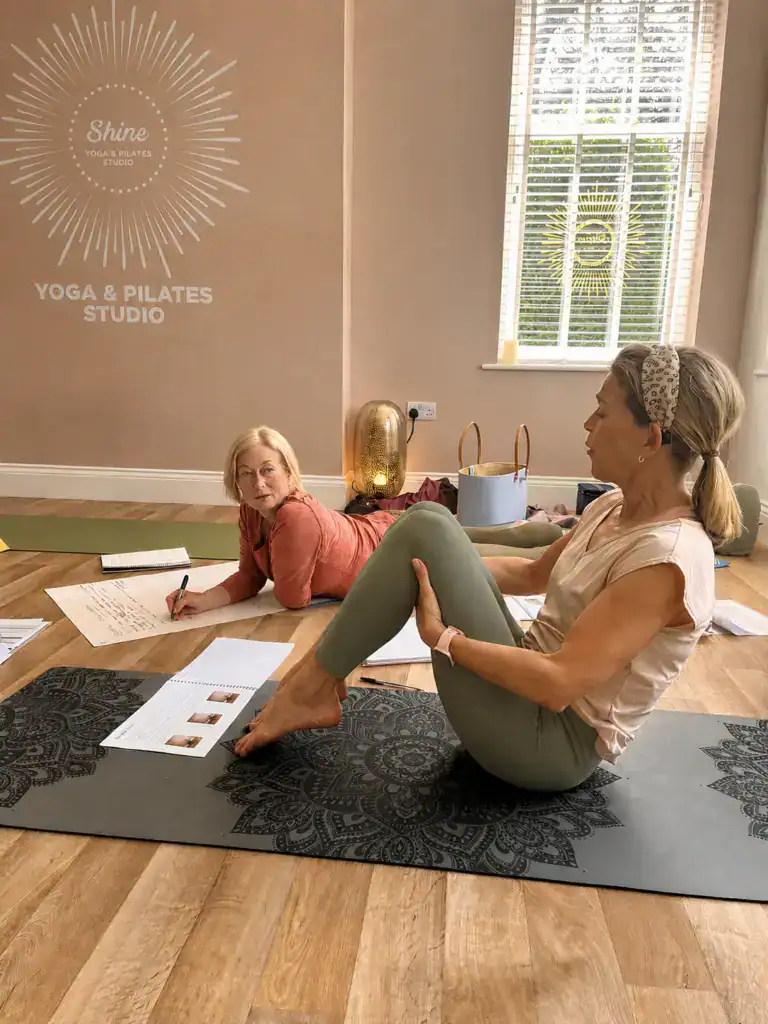 Two women sitting and laying on the floor of the yoga studio at Shine at The Hollies working in the Pilates Teacher Training course