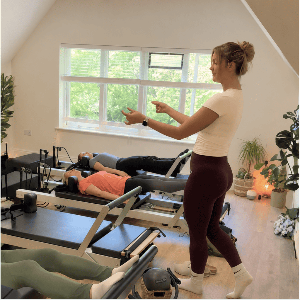 group of ladies laying on Reformer Pilates machines while the instructor stands and guides them in their movements at a reformer pilates class at Shine at the Holiies