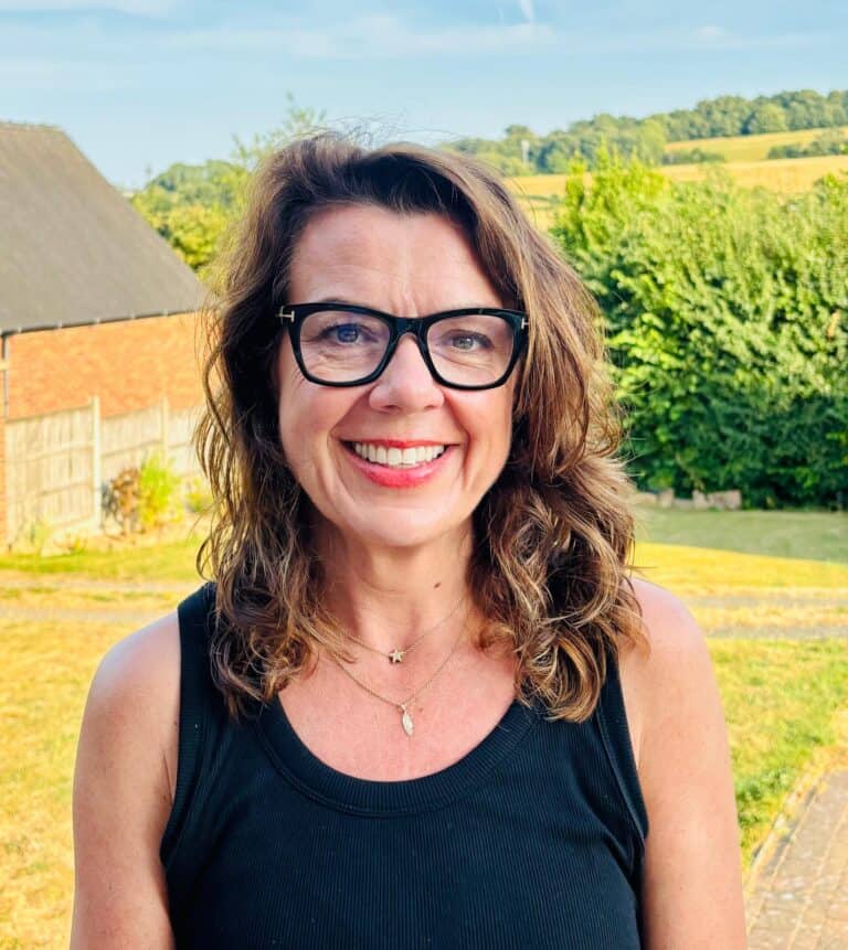 Lisa Langhorn, Co-Owner of Shine at the Hollies. Woman with brown hair, and glasses, smiling, standing in the countryside on a summers day