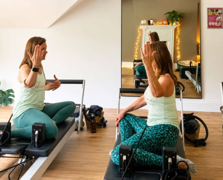 Tabitha Wright owner of Shine at The Hollies is sitting on a Reformer Pilates machine demonstrating an exercise to another woman who is sitting on another Reformer Pilates machine in the Reformer Pilates Teacher Training course