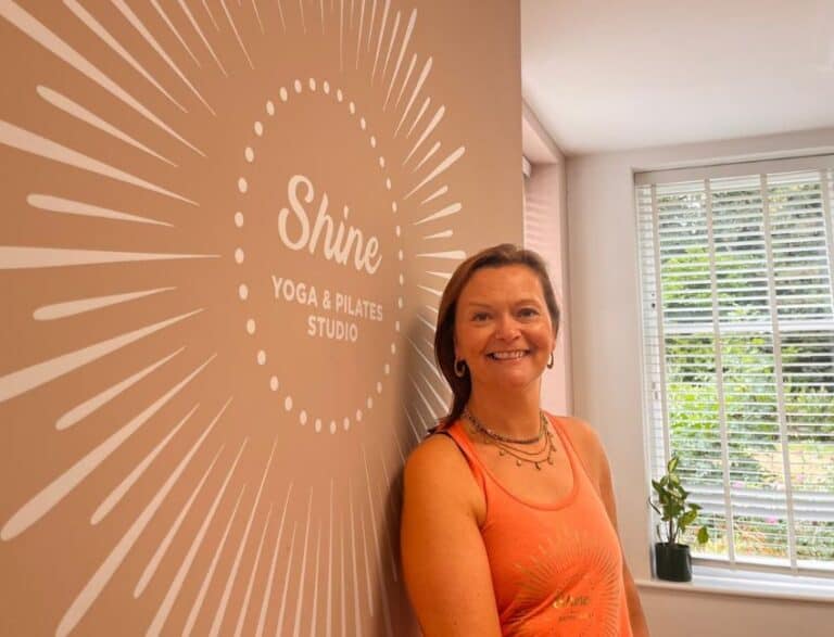 Tabitha Wright owner of Shine at The Hollies leaning on the wall of the yoga studio in front of the shine logo